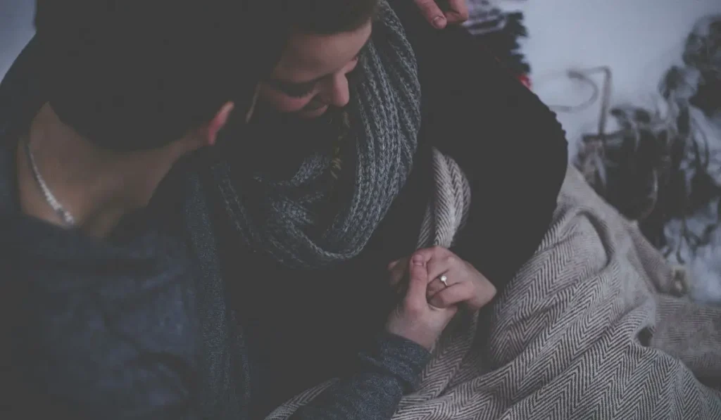 Couple holding hands, showcasing an engagement ring, wrapped in a cozy blanket during a winter engagement moment.