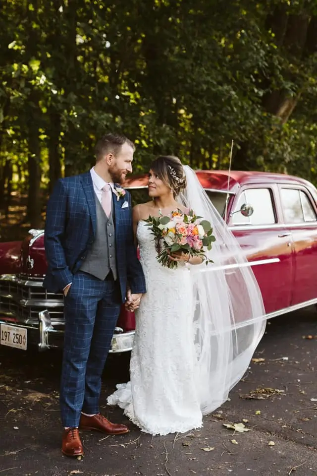 Bride and groom posing by vintage red car, showcasing elegant and memorable wedding transportation options.