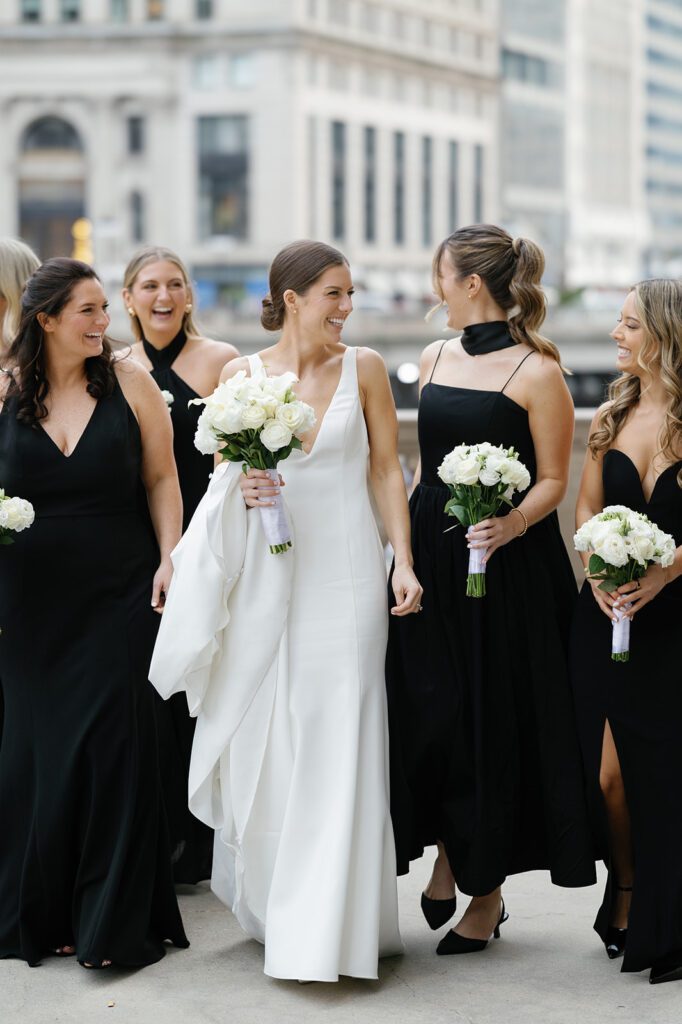 Bride in white gown with bouquet surrounded by bridesmaids in black dresses, smiling together in urban Chicago setting with tall buildings behind