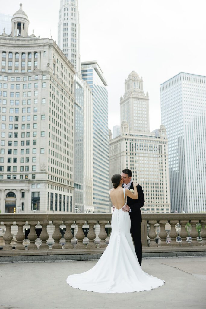Bride in low-back gown and groom in tuxedo embrace on stone terrace with Chicago skyline in background, capturing a romantic editorial wedding moment