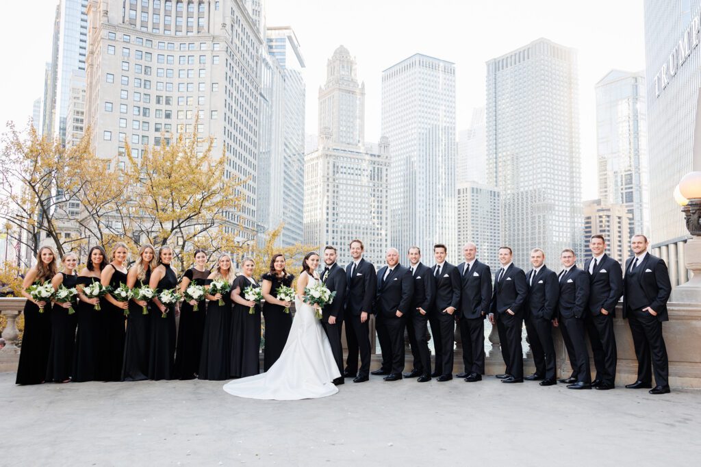 Bride and groom pose with wedding party in formal black attire beneath autumn trees and Chicago skyscrapers, including the Trump Tower, capturing a classic city celebration with editorial style