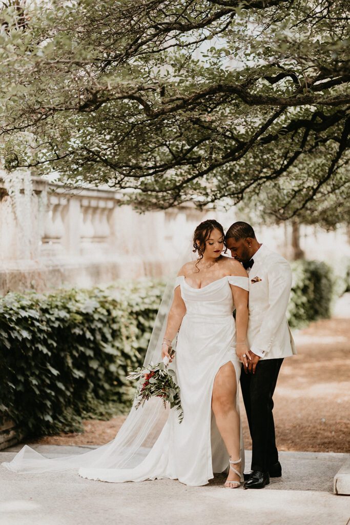 Couple standing beneath lush tree canopy in formal wedding attire, surrounded by ivy and stone — a serene moment in their wedding planning timeline.