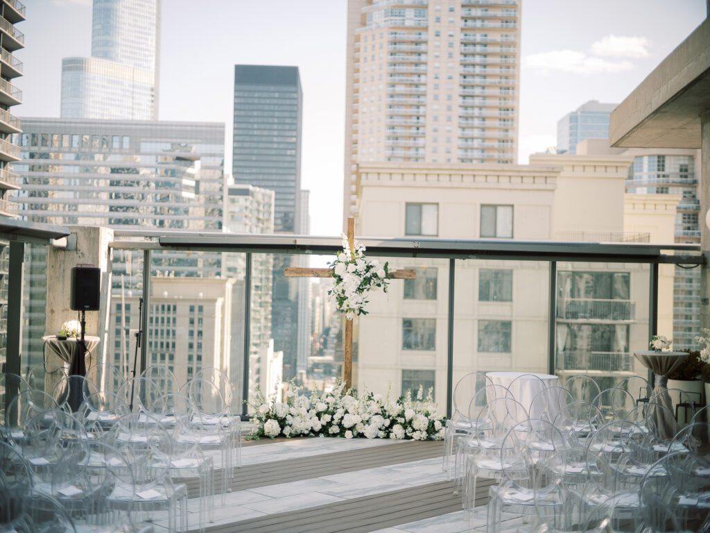 Rooftop wedding ceremony setup with floral cross, transparent chairs, and city backdrop