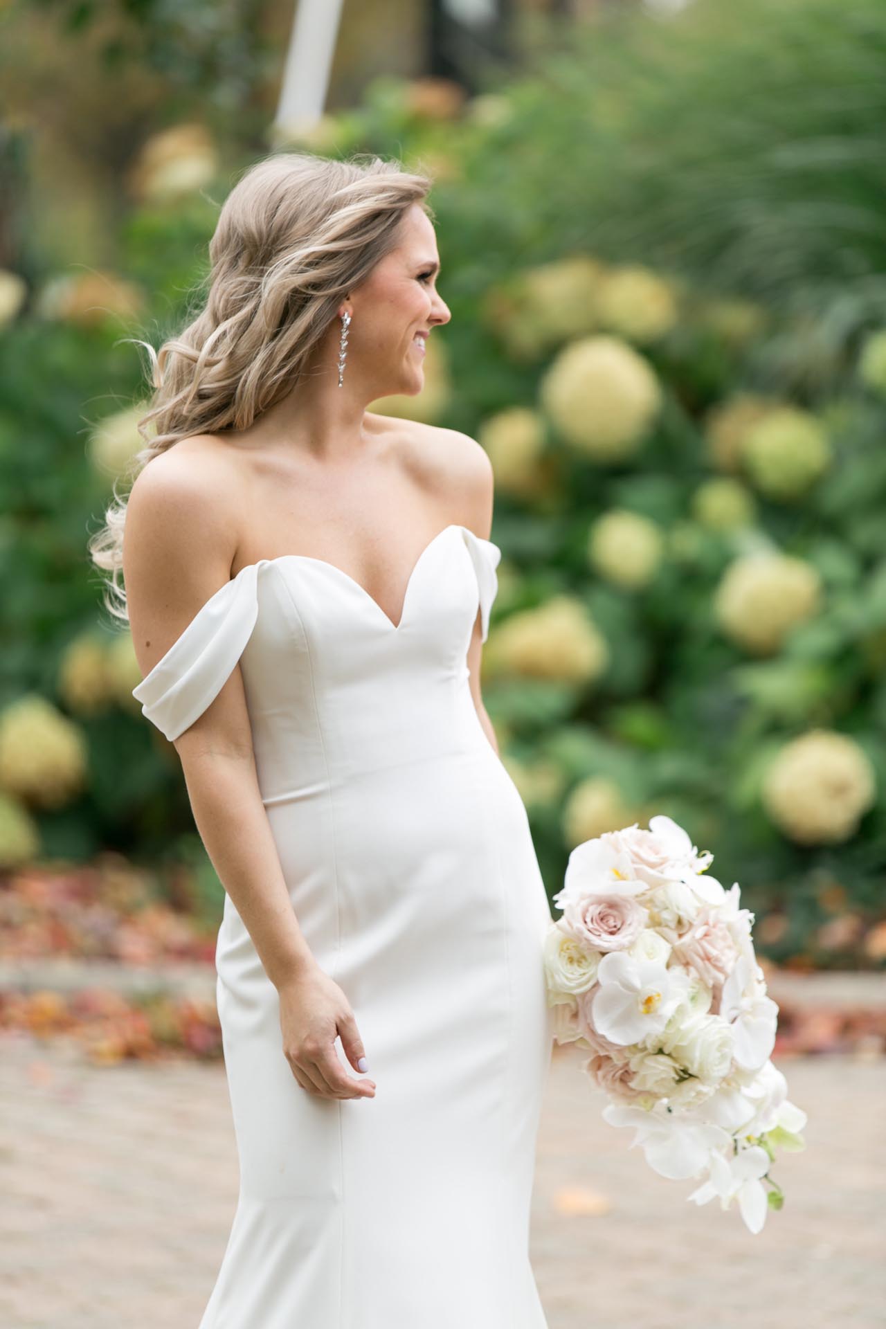 Bride in off‑shoulder gown holding white and blush bouquet on garden path in Chicago.