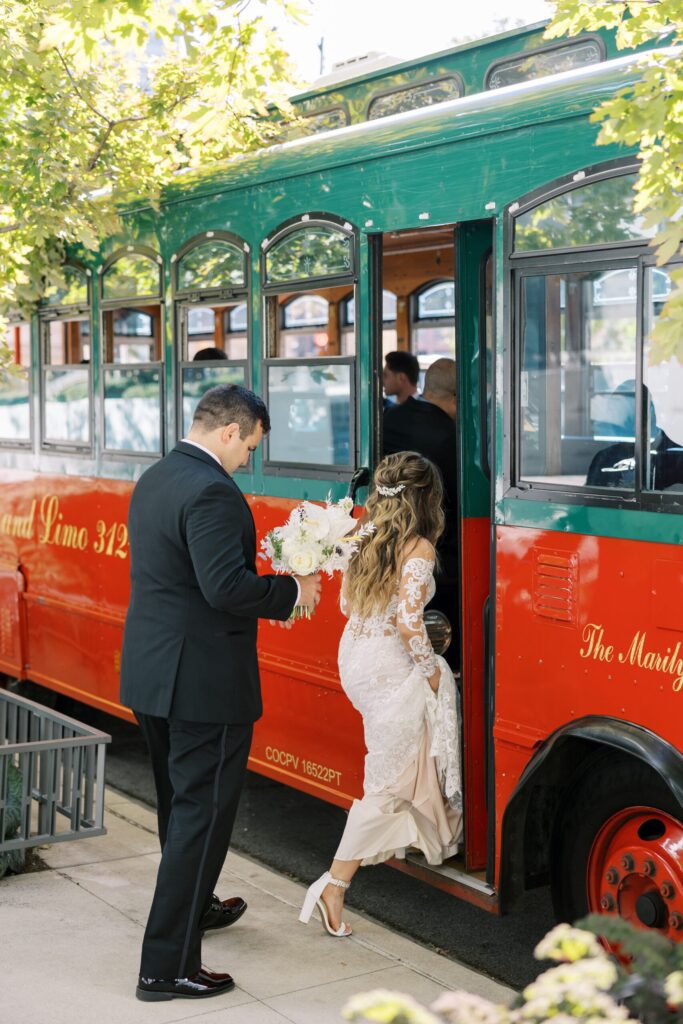 Bride and groom board “The Marilyn” red-and-green vintage trolley with white florals in hand, surrounded by greenery and formally dressed guests.