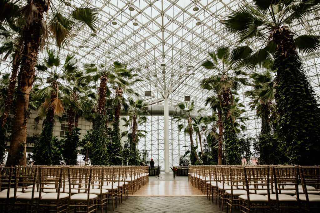 Indoor wedding ceremony at Navy Pier featuring a glass ceiling, tropical greenery, and gold chairs lining a central aisle flooded with natural light.