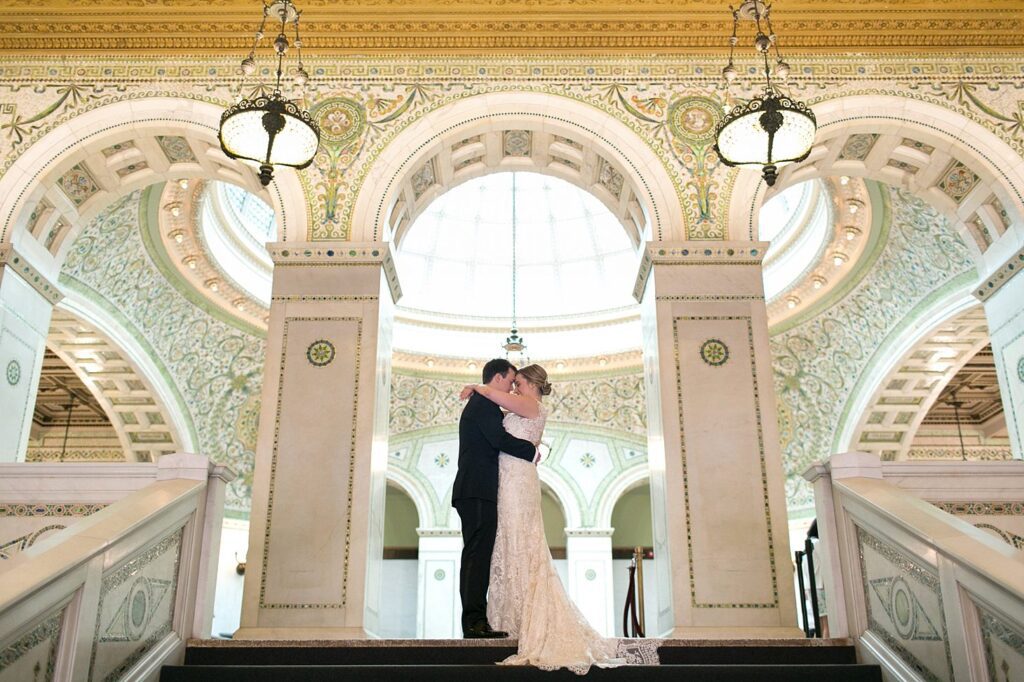 Wedding couple sharing a kiss at the top of a grand staircase beneath mosaic arches and a domed skylight at the Chicago Cultural Center.