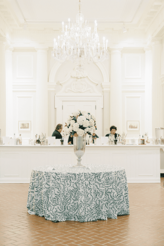 Wedding escort card table at the Chicago History Museum featuring a tall floral centerpiece, classical architecture, and a chandelier-lit bar backdrop.