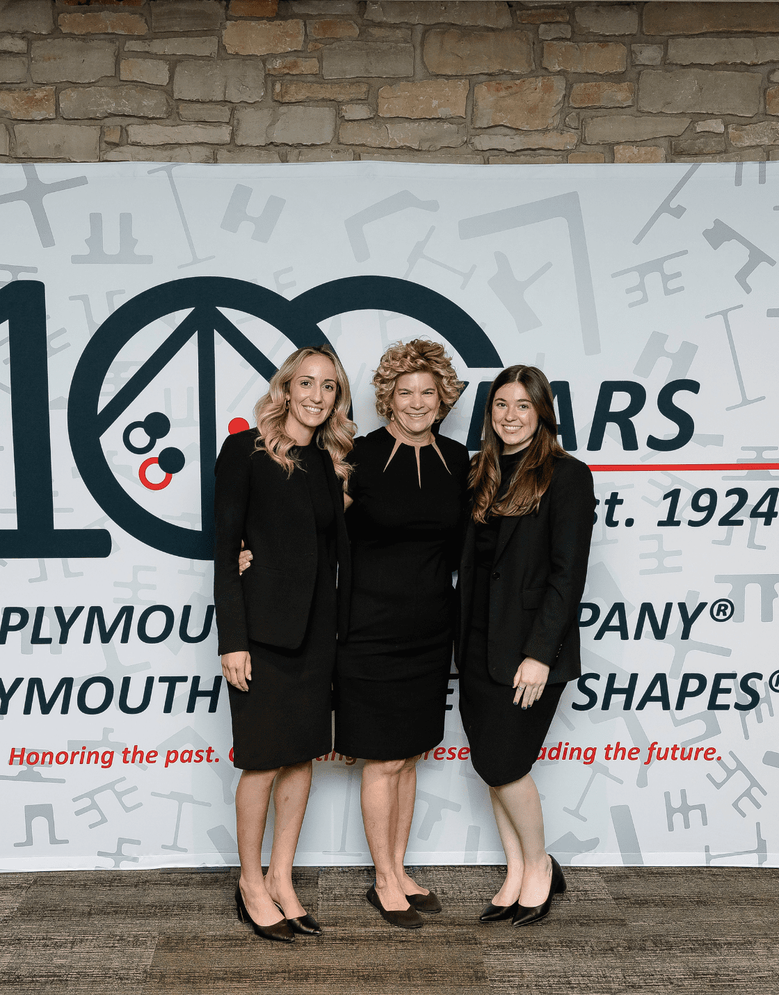 Three individuals in formal attire pose in front of Plymouth Tube Company’s 100-year celebration backdrop at a destination corporate event.