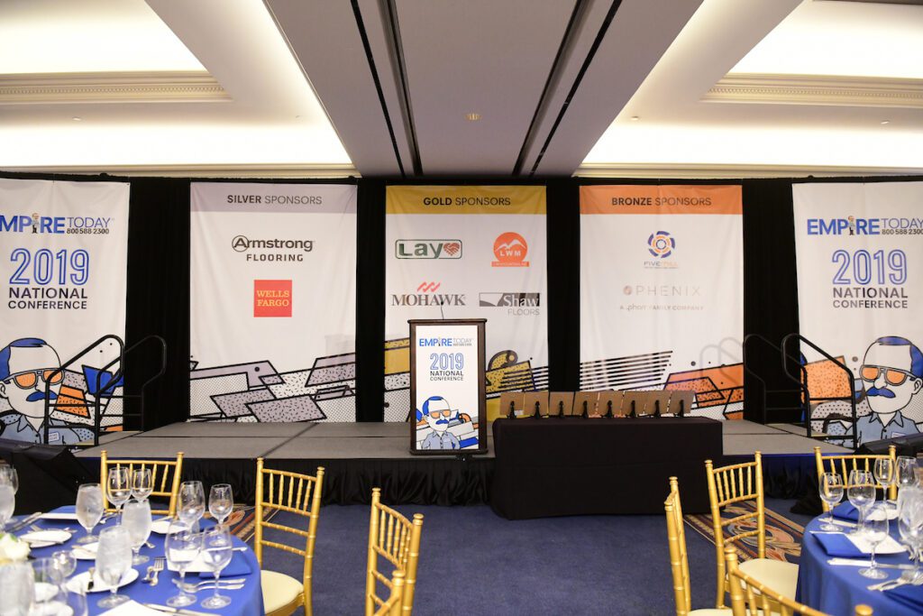 Branded stage setup for Empire Today’s national conference with sponsor banners and dining tables in foreground.