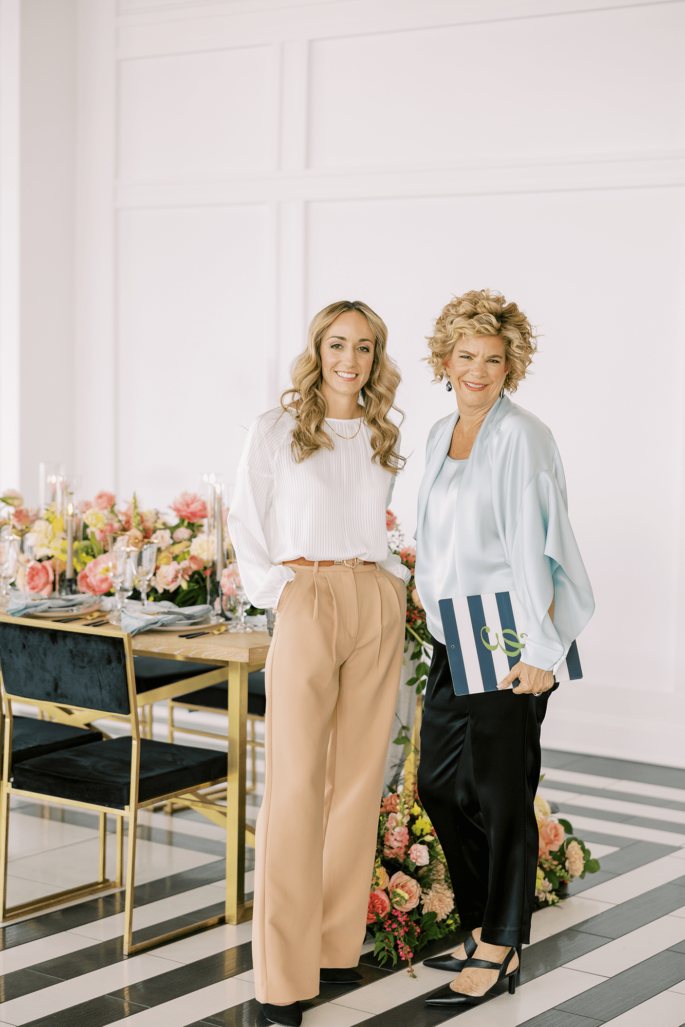 Two event planners stand beside an elegantly styled dining table with florals and candles, captured in a polished, editorial setting during event setup.
