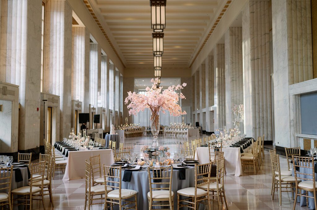 Elegant wedding reception in Chicago’s Historic Lobby featuring cherry blossom centerpieces, gold chairs, and marble columns under geometric chandeliers.