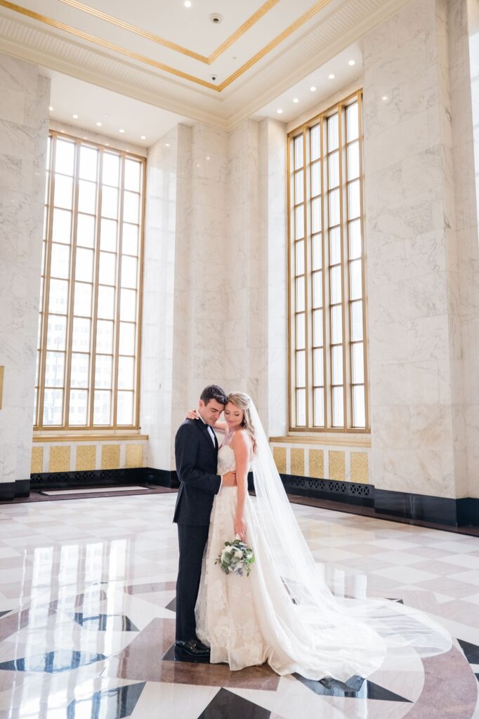 Bride and groom pose in the Historic Lobby, surrounded by marble columns and golden window trim.