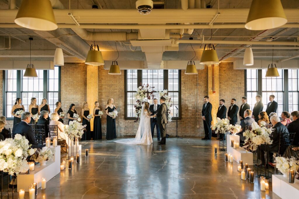 Romantic indoor wedding ceremony at the Old Post Office with a floral arch, black-clad bridal party, and candle-lined aisle in an industrial-chic space.