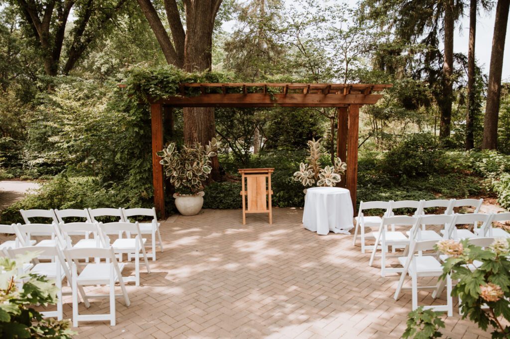 Intimate Morton Arboretum venue space for a ceremony under a vine‑covered pergola with white chairs and lush greenery.