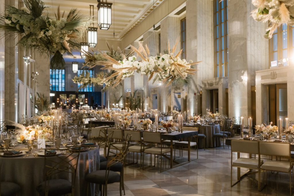 Dramatic suspended pampas and white floral installation above a long reception table in the Old Post Office’s marble-columned hall, flanked by chevron bar counters.