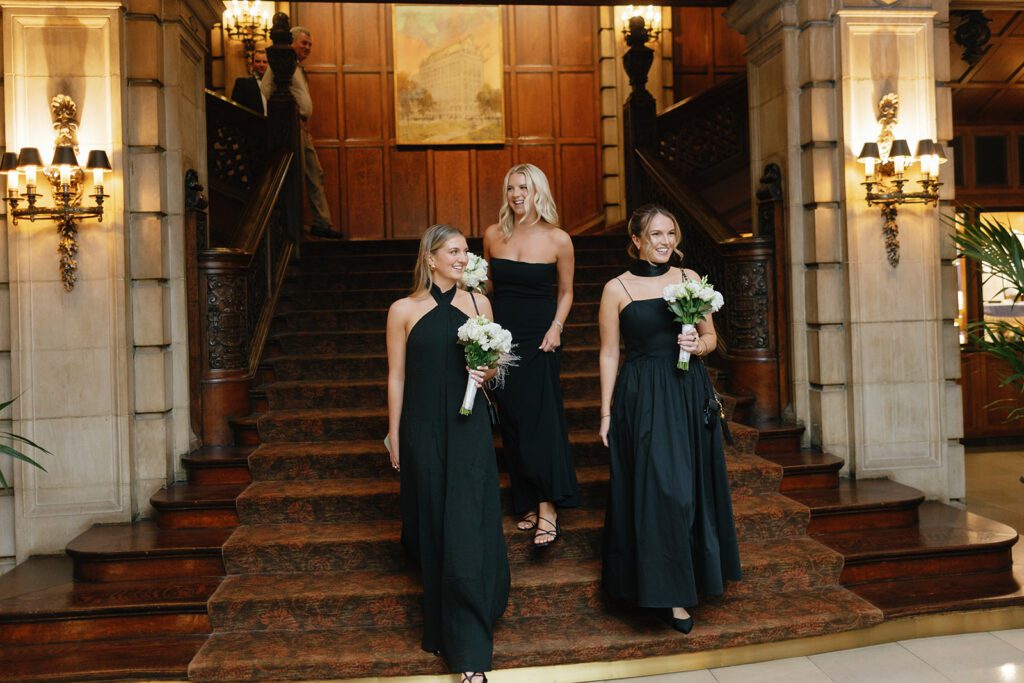 Three women in black gowns descending a grand staircase in an upscale venue, showcasing elegant black tie wedding attire.