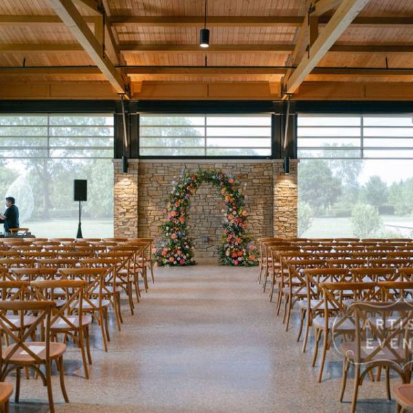 Indoor wedding ceremony at firefly pavilion with wood beams, floral arch, and natural light from large windows.